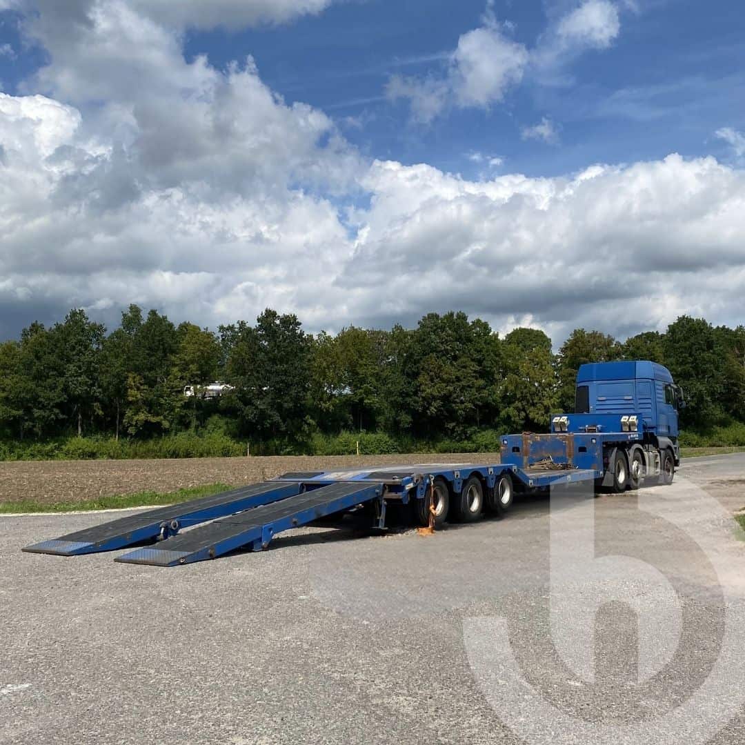 Blaue Zugmaschine mit blauem Tieflader in Rückansicht auf offenem Gelände.